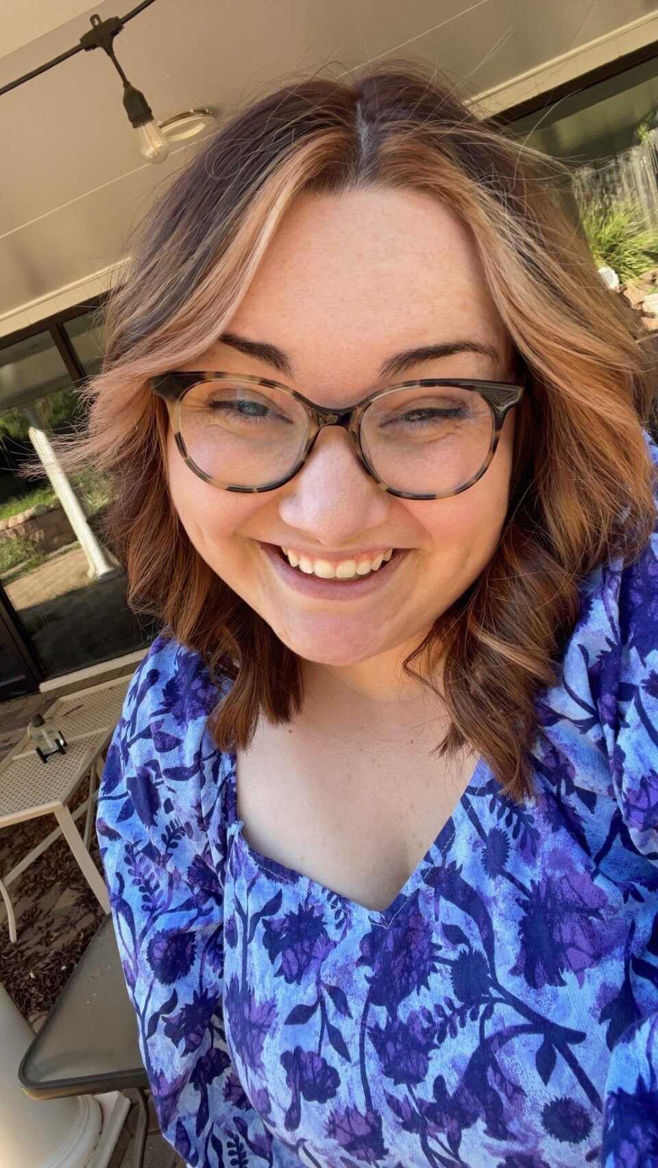 Smiling woman with wavy brown hair, glasses, and a purple floral top in an outdoor patio setting.
